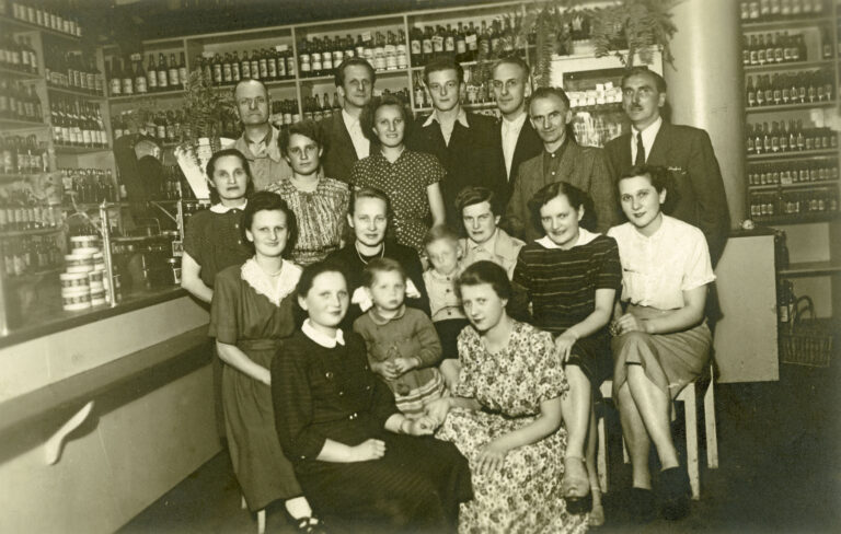 Personel we wnętrzu sklepu spożywczego w Bydgoszczy fotografia zbiorowa kilkanaście kobiet i mężczyzn w średnim i młodym wieku, część siedzi na krzesłach dwie małe dziewczynki z tyłu lada sklepowa za nią półki do sufitu z butelkami
