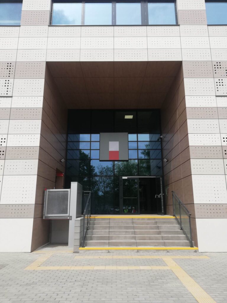 Wejście główne do budynku, schody obok podnośnik do wózków nad otwartymi drzwiami logo archiwów państwowych