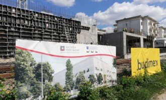 Prace na poziomie pierwszym i drugim, widok od strony wejścia głównego Prace na poziomie pierwszym i drugim, widok od strony wejścia głównego, ściana parteru w budowie za płotem na którym wiszą banery Archiwum Państwowego z wizualizacją przyszłego budynku i drugi żółty z czarnym logo firmy Budimex