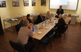 Osiem osób przy stole, mężczyźni i kobiety siedzą przy stole dookoła przemawia do nich dyrektor Archiwum ubrany w garnitur, na stole woda w małych butelkach i ciasto na talerzach w tle gabloty wystawowe na ścianach plansze z fotografiami