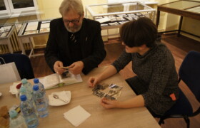 Siwy mężczyzna z brodą w garniturze i kobieta siedzą przy stole i przeglądają lużne czarno-białe fotografie w tle gabloty wystawowe z archiwaliami