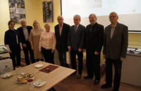 Osiem osób w tym trzy kobiety wszyscy elegancko ubrani stoją w rzędzie w tle gabloty wystawowe fragment ekranu na ścianie także plansze ze zdjęciami wnętrza archiwum