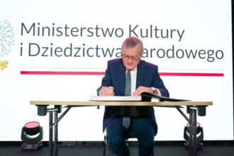 Minister Gliński dokonuje wpisu do księgi pamiątkowej siedzi na tle logo Ministerstwa Kultury