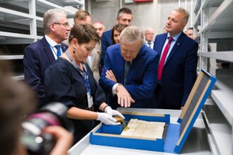 W archiwalnym skarbcu, minister ogląda średniowieczny dokument, pracowniczka prezentuje pieczęć woskową w tle uczestnicy wydarzenia
